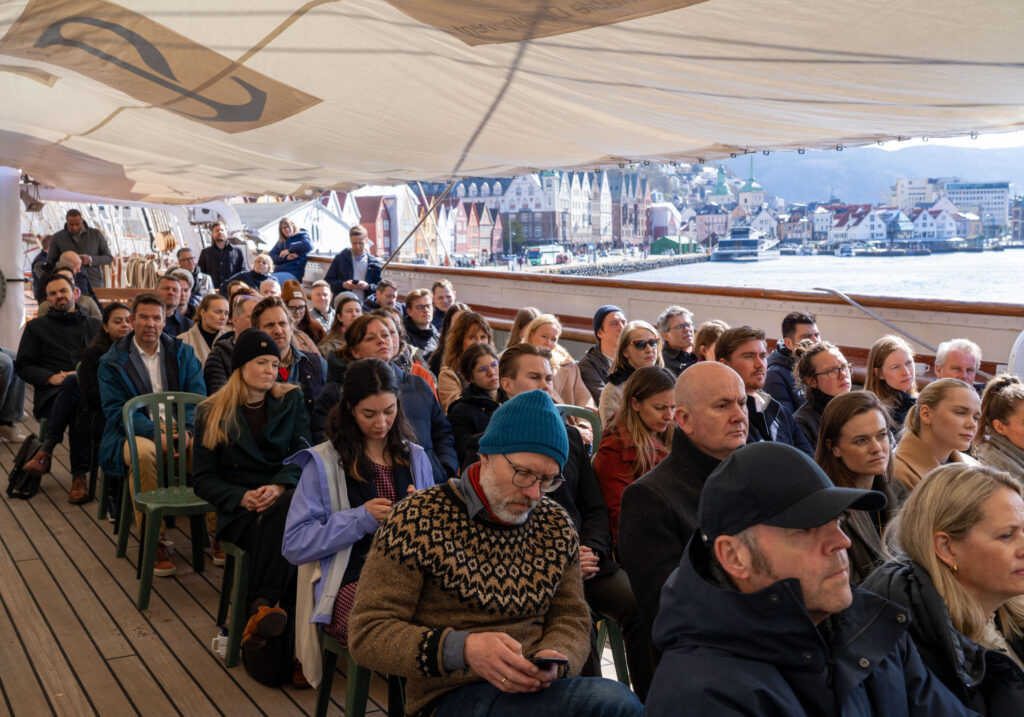 Over 50 mennesker sitter på stoler på dekket til et skip og ser på et foredrag. Man ser Bryggen og Vågen i Bergen i sollys i bakgrunnen.