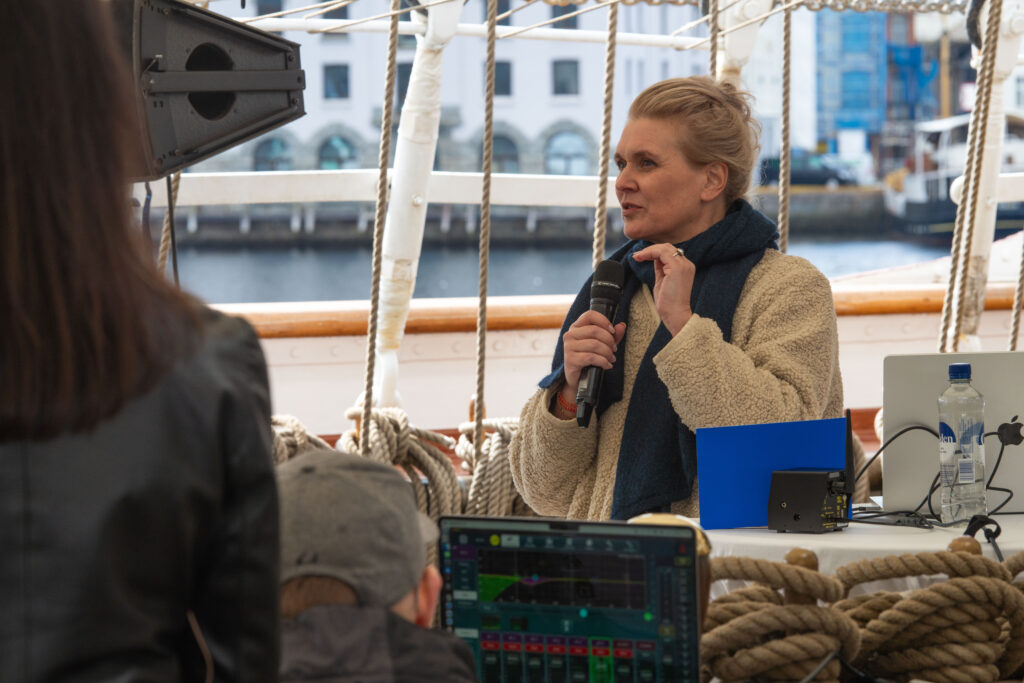 En kvinne snakker til en forsamling i en mikrofon. Hun står på dekket til skipet Statsraad Lehmkuhl. 